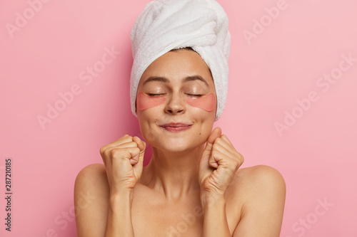 Spa woman makes mask for hair, applies hydrogel patches, clenches fists as anticipates for cosmetic product effect, stands shirtless with eyes closed, isolated over pink background. Eye skin care