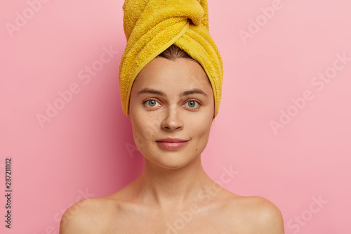 Close up shot of gorgeous fresh European woman with towel on head  has clean face  healthy skin  stands shirtless  takes shower  going to apply makeup  has natural beauty. Body care concept.