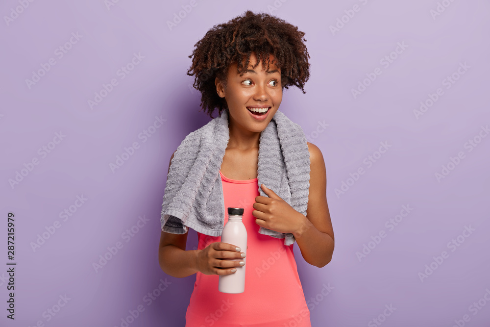 Photo of delighted dark skinned sportswoman has Afro haircut, looks aside with smile, dressed in pink top, carries bottle, drinks water while feels thirsty during training, excercises indoor. Photo of delighted dark skinned sportswoman has Afro haircut, looks aside with smile, dressed in pink top, carries bottle, drinks water while feels thirsty during training, excercises indoor.