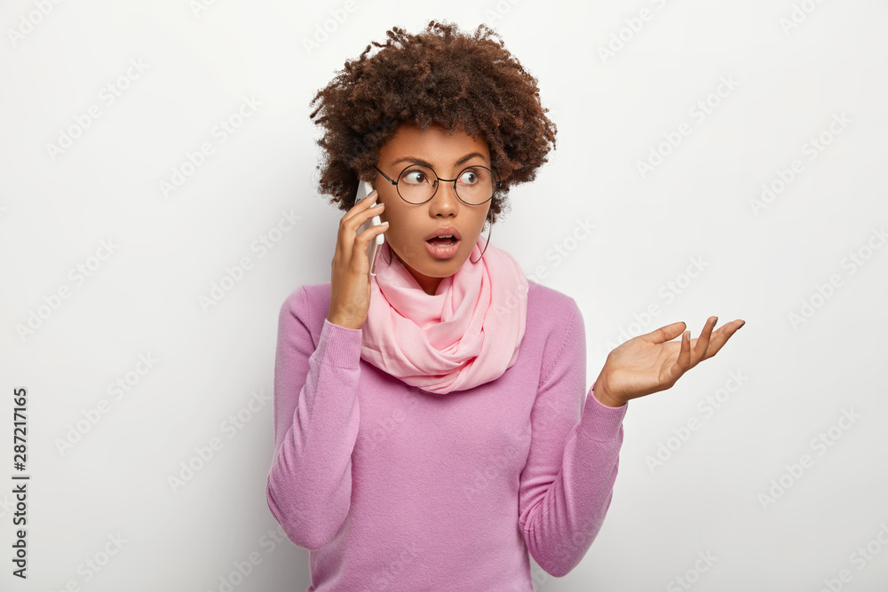 Puzzled female with Afro hairstyle, has conversation with colleague or friend, tries to solve problem via smartphone, focused aside, wears optical glasses, purple casual jumper and scarf around neck Puzzled female with Afro hairstyle, has conversation with colleague or friend, tries to solve problem via smartphone, focused aside, wears optical glasses, purple casual jumper and scarf around neck