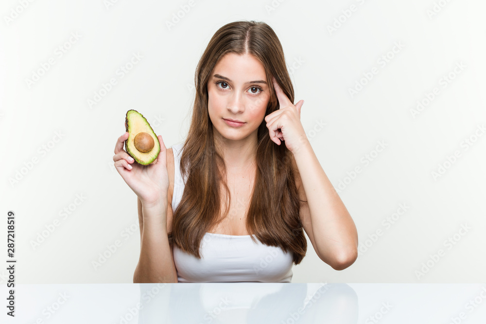 Young caucasian woman holding an avocado pointing his temple with finger, thinking, focused on a task. Young caucasian woman holding an avocado pointing his temple with finger, thinking, focused on a task.