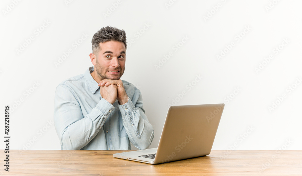 Young handsome man working with his laptop keeps hands under chin, is looking happily aside. Young handsome man working with his laptop keeps hands under chin, is looking happily aside.