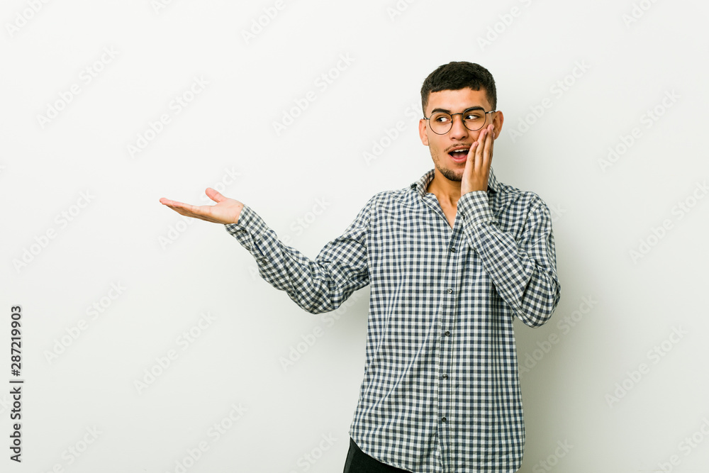Young hispanic man holds copy space on a palm, keep hand over cheek. Amazed and delighted.