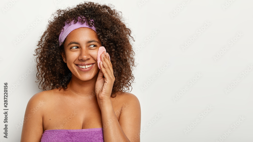 Close up portrait of cheerful African American woman with curly bushy hair, wears purple headband, wrapped in towel, wipes skin with cosmetic sponge, isolated over white wall with copy space Close up portrait of cheerful African American woman with curly bushy hair, wears purple headband, wrapped in towel, wipes skin with cosmetic sponge, isolated over white wall with copy space