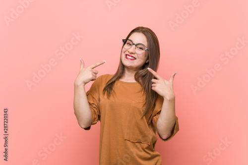 Young student woman wearing eyeglasses smiles, pointing fingers at mouth.