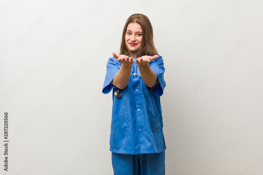 Young nurse woman against a white wall holding something with palms, offering to camera. Young nurse woman against a white wall holding something with palms, offering to camera.