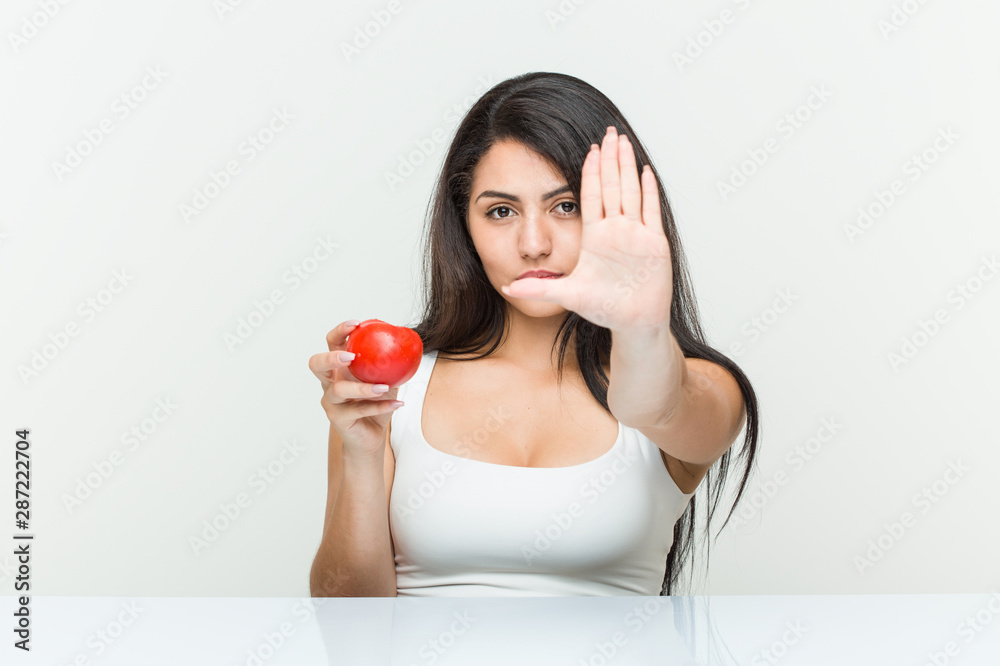 Young hispanic woman holding a tomato standing with outstretched hand showing stop sign, preventing you. Young hispanic woman holding a tomato standing with outstretched hand showing stop sign, preventing you.