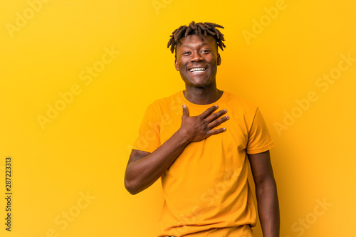 Young black man wearing rastas over yellow background laughs out loudly keeping hand on chest.