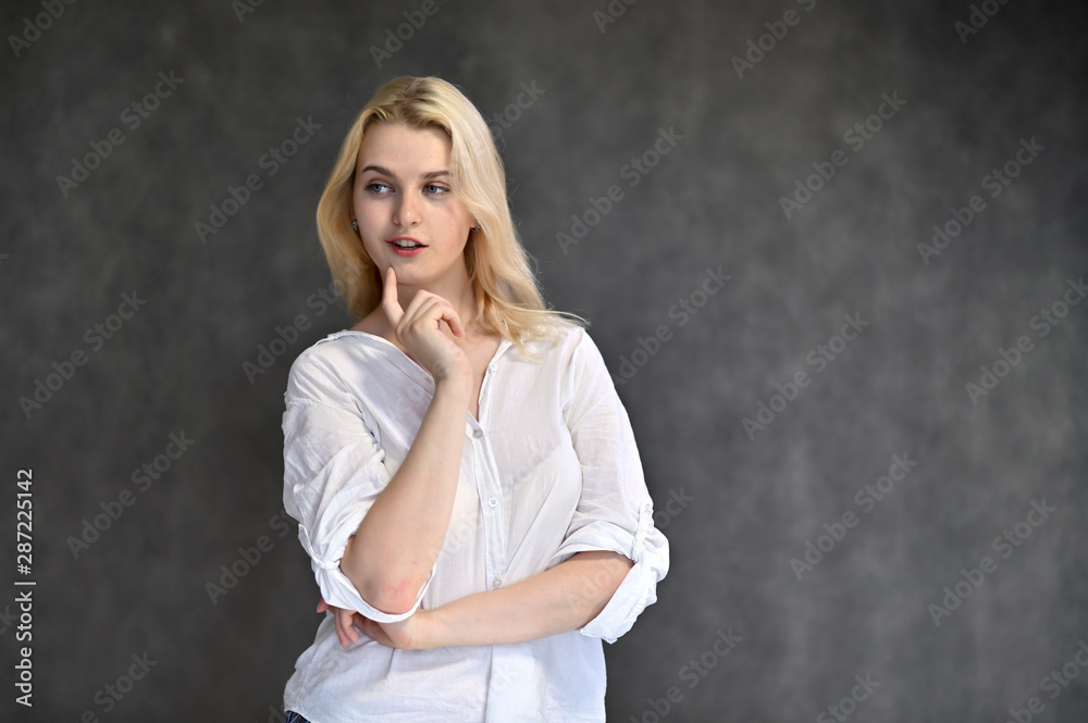 Portrait of a cute blonde girl, a young woman in a white shirt with beautiful curly hair on a gray background. Shows hands to the sides. Beauty, brightness, emotions. Portrait of a cute blonde girl, a young woman in a white shirt with beautiful curly hair on a gray background. Shows hands to the sides. Beauty, brightness, emotions.