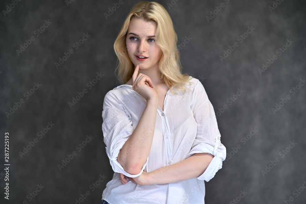 Portrait of a cute blonde girl, a young woman in a white shirt with beautiful curly hair on a gray background. Shows hands to the sides. Beauty, brightness, emotions. Portrait of a cute blonde girl, a young woman in a white shirt with beautiful curly hair on a gray background. Shows hands to the sides. Beauty, brightness, emotions.