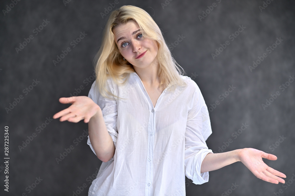 Portrait of a cute blonde girl, a young woman in a white shirt with beautiful curly hair on a gray background. Shows hands to the sides. Beauty, brightness, emotions. Portrait of a cute blonde girl, a young woman in a white shirt with beautiful curly hair on a gray background. Shows hands to the sides. Beauty, brightness, emotions.
