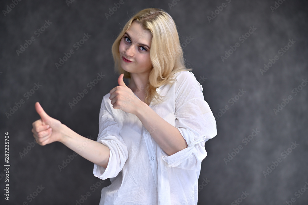 Portrait of a cute blonde girl, a young woman in a white shirt with beautiful curly hair on a gray background. Shows hands to the sides. Beauty, brightness, emotions. Portrait of a cute blonde girl, a young woman in a white shirt with beautiful curly hair on a gray background. Shows hands to the sides. Beauty, brightness, emotions.