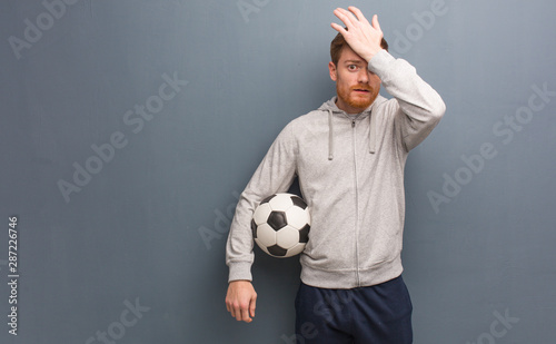 Young redhead fitness man worried and overwhelmed. He is holding a soccer ball.