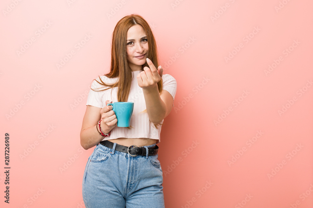 Young caucasian woman holding a cup pointing with finger at you as if inviting come closer. Young caucasian woman holding a cup pointing with finger at you as if inviting come closer.