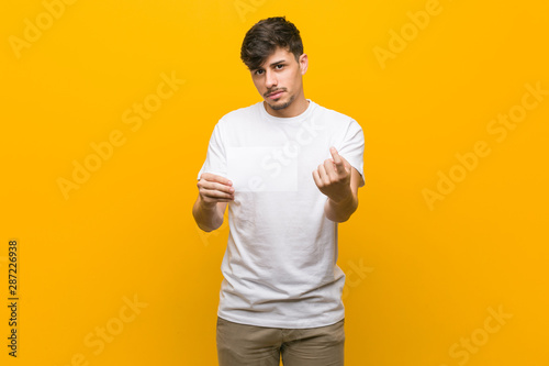 Young hispanic man holding a placard pointing with finger at you as if inviting come closer.