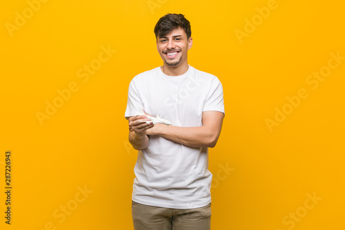 Young hispanic man holding an airplane icon smiling confident with crossed arms.