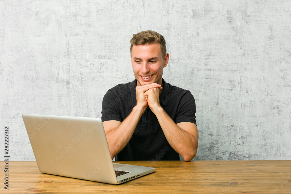 Young entrepreneur working with his laptop on a desk keeps hands under chin, is looking happily aside. Young entrepreneur working with his laptop on a desk keeps hands under chin, is looking happily aside.