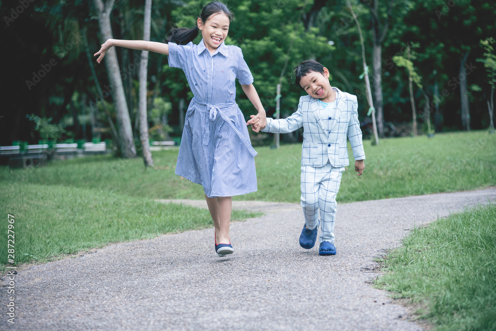 Asian children, sisters and brothers held hands walked on the streets in the park Are smiling, fun and happy with blurred soft of green trees background, to children and family concept. Asian children, sisters and brothers held hands walked on the streets in the park Are smiling, fun and happy with blurred soft of green trees background, to children and family concept.