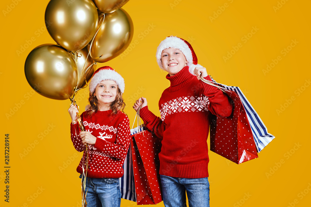 Cute children in red Santa hats and sweaters hugging and holding shopping bags and golden balloons on yellow background