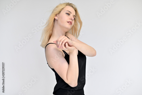 Portrait of a cute blonde student girl, a young woman in a black dress with beautiful curly hair on a white background in different poses. Shows hands to the sides. Beauty, brightness, emotions.