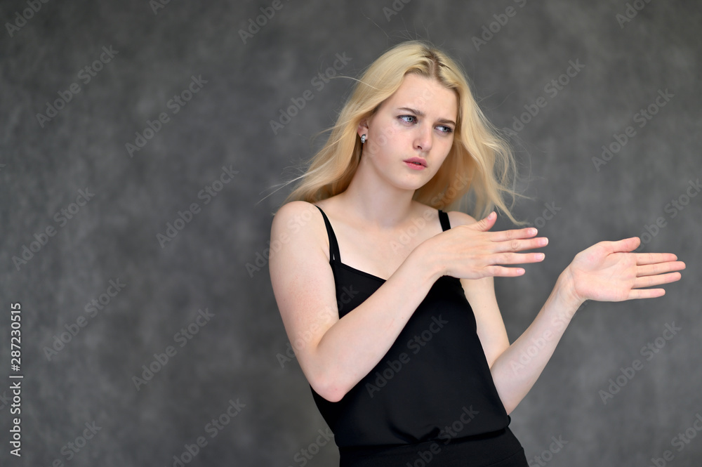 Portrait of a cute blonde student girl, a young woman in a black dress with beautiful curly hair on a gray background in different poses. Shows hands to the sides. Beauty, brightness, emotions. Portrait of a cute blonde student girl, a young woman in a black dress with beautiful curly hair on a gray background in different poses. Shows hands to the sides. Beauty, brightness, emotions.