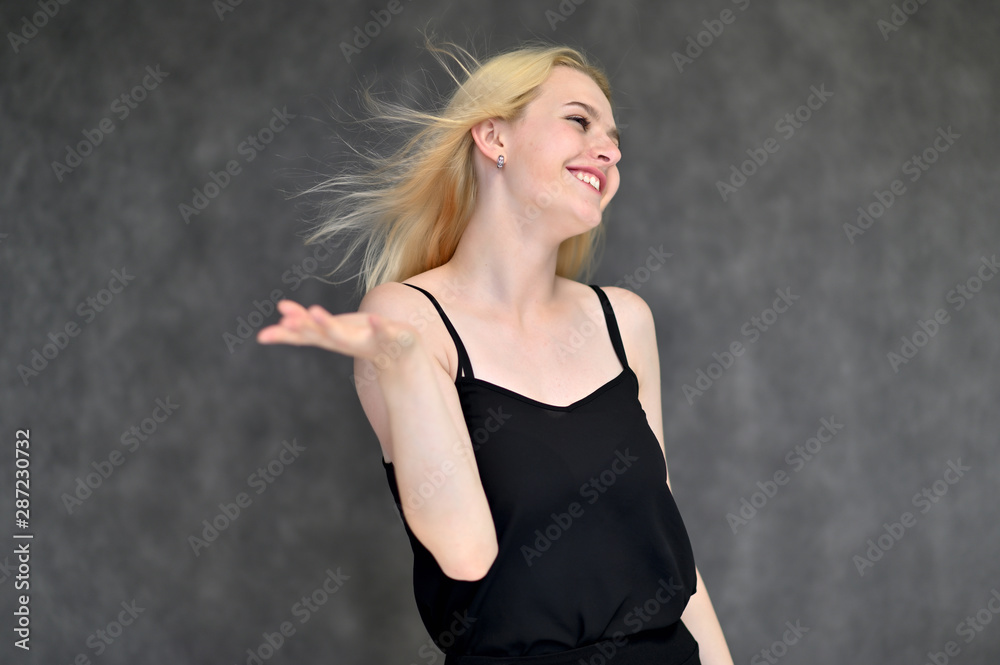 Portrait of a cute blonde student girl, a young woman in a black dress with beautiful curly hair on a gray background in different poses. Shows hands to the sides. Beauty, brightness, emotions. Portrait of a cute blonde student girl, a young woman in a black dress with beautiful curly hair on a gray background in different poses. Shows hands to the sides. Beauty, brightness, emotions.