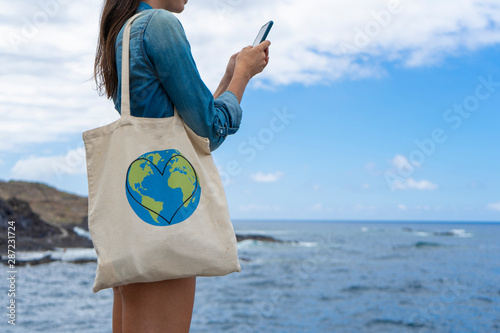 Girl holding cotton canvas while typing with her cellphone in the sea. Conscious Woman using ecology carrier handbag and messaging, smiling. Teenager with organic sack