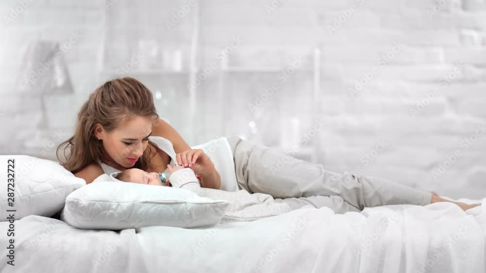 Lovely smiling mother lying on comfortable white bed lulling cute baby having positive emotion Lovely smiling mother lying on comfortable white bed lulling cute baby having positive emotion