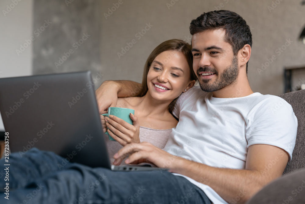Smiling young couple relaxing on a couch and drinking tea at home while using laptop computer Smiling young couple relaxing on a couch and drinking tea at home while using laptop computer
