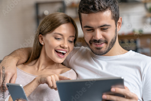 Close up shot of young happy couple hugging and using smartphone, digital tablet while sitting on sofa at home