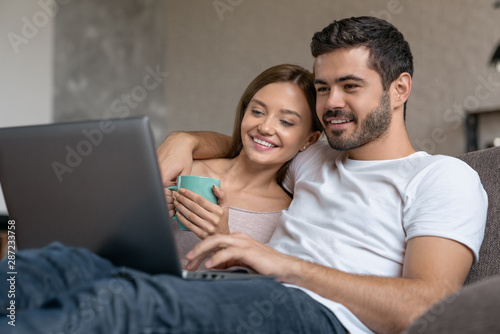 Smiling young couple relaxing on a couch and drinking tea at home while using laptop computer