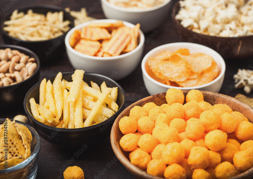 All classic potato snacks with peanuts, popcorn and onion rings and salted pretzels in bowl plates on wooden background. Twirls with sticks and potato chips and crisps with nachos and cheese balls. All classic potato snacks with peanuts, popcorn and onion rings and salted pretzels in bowl plates on wooden background. Twirls with sticks and potato chips and crisps with nachos and cheese balls.