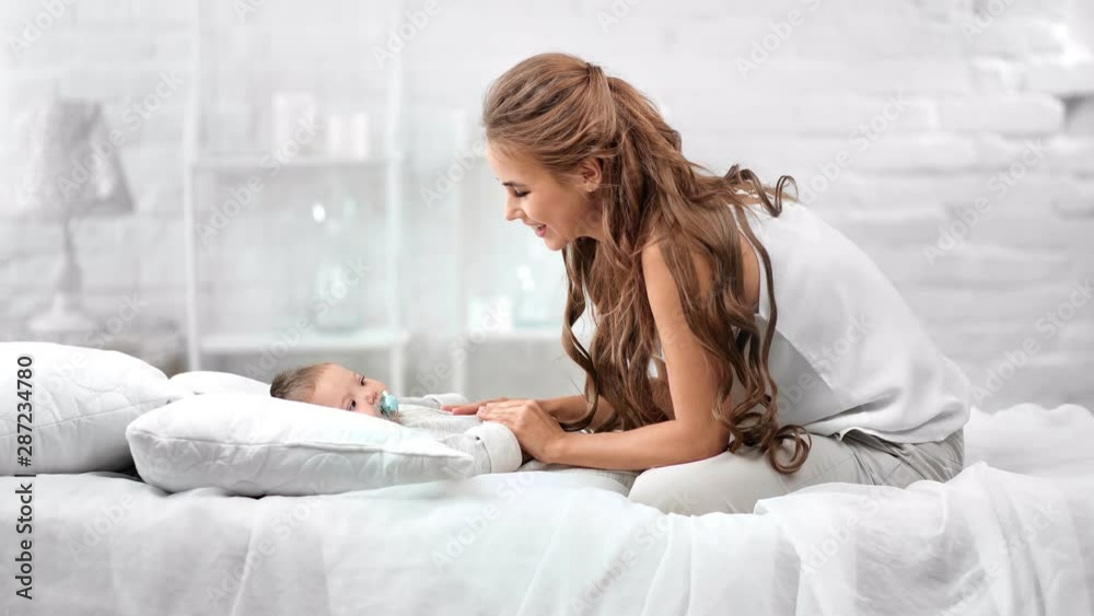 Smiling mother enjoying caress cute baby resting on bed surrounded by white room interior Smiling mother enjoying caress cute baby resting on bed surrounded by white room interior