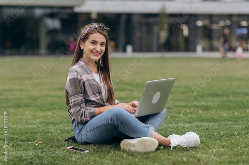 Pretty young woman sitting on green grass in park with legs crossed during summer day while using laptop