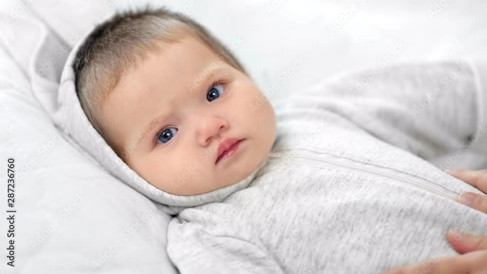 Focused face of beautiful little baby lying on bed during caress by female hand looking at camera Focused face of beautiful little baby lying on bed during caress by female hand looking at camera
