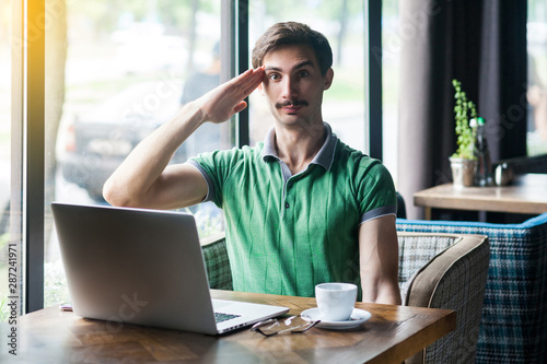 Foto Yes sir! Young serious businessman in green t-shirt sitting and looking at camera with salute gesture and ready to complete the mission