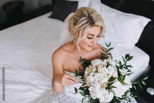 Beautiful blonde bride is sitting on the bed and looking at a bouquet of roses. The morning of the bride. Wedding closeup portrait of a cute girl in a hotel. Concept and photography.