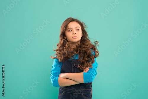 Awesome beauty. Adorable baby girl keeping arms crossed on blue background. Brunette girl with long wavy hair wearing casual style. Cute little girl of fashion. Small girl with beauty look