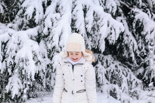 Portrait of little girl in winter. Toddler playing with snow in park. Child in white warm winter clothes and knit hat looking at the falling snow In winter forest. Snowfall. Winter activities for kids
