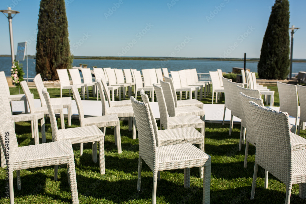 Fototapeta premium Beautiful outgoing wedding set up.Romantic wedding ceremony , wedding outdoor on the lawn water view. Wedding decor. White wooden chairs on a green lawn. White armchairs for guests