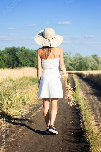 Young beautiful woman in a white dress and hat holds a bouquet with wildflowers on the road between the milky field. Concept of outdoor recreation, a trip to the village © Alex