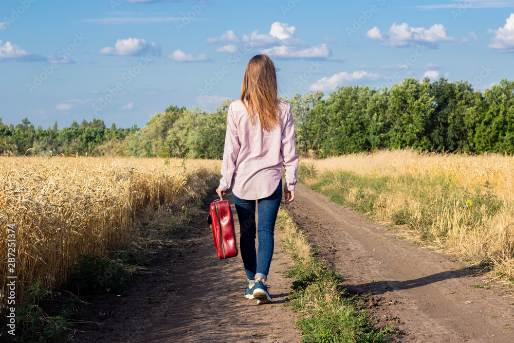 Beautiful young woman with a suitcase is walking along a road between wheat fields in the background. Travel concept, vacation