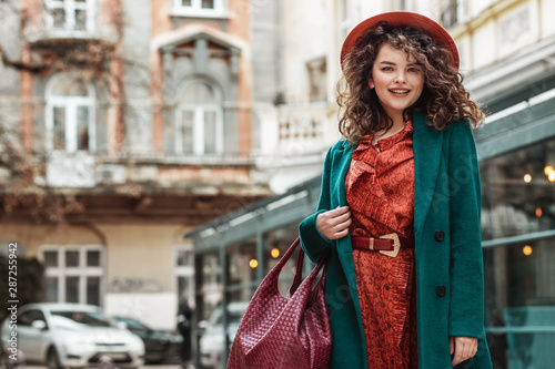Outdoor portrait of young happy smiling fashionable lady wearing trendy orange dress, hat, belt, green classic coat, holding red handbag, posing in street of European city. Copy, empty space for text © Victoria Fox