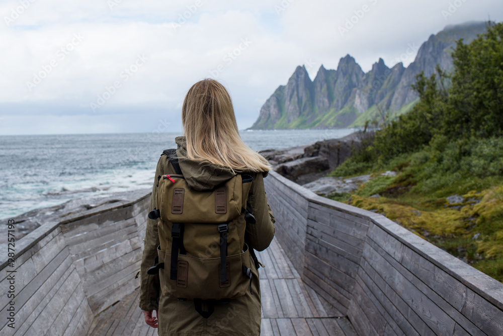 Blond hair girl with backpack enjoy beautiful nature landscape in North. Ocean and mountain. Tourist landmark in Norway. Amazing scenic outdoors view. Travel, adventure, lifestyle. Lofoten Islands