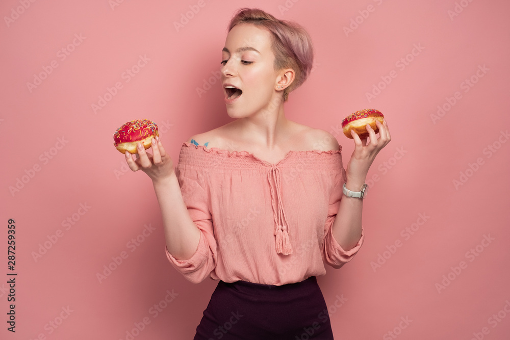 A girl with short pink hair, holds donuts and opens her mouth, reaches for one of them, on a pink background. A girl with short pink hair, holds donuts and opens her mouth, reaches for one of them, on a pink background.