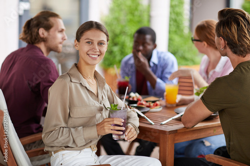 Portrait of pretty young woman smiling at camera and holding cocktail while enjoying lunch together with friends in cafe, copy space