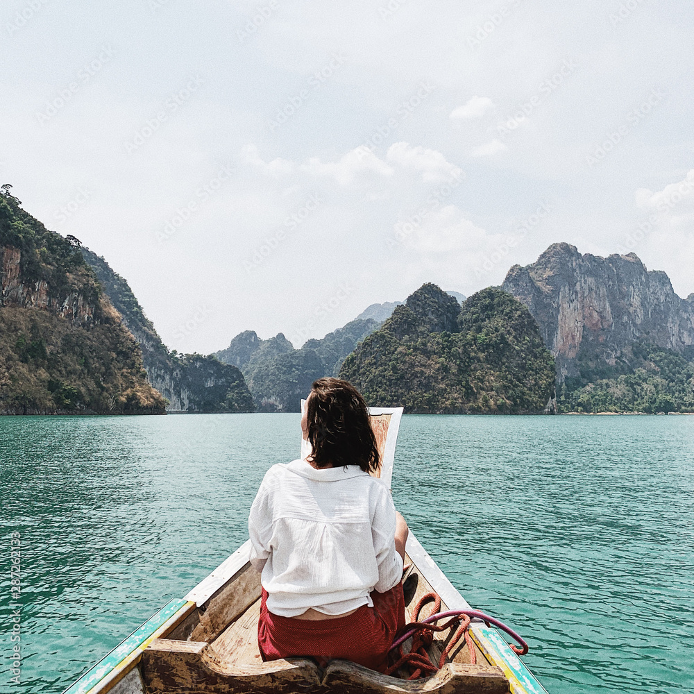 Young woman in red skirt and white blouse sitting on wooden boat watching at exotic and tropical dark green big islands with rocks and turquoise lake in Thailand. Travel holiday and adventure concept.