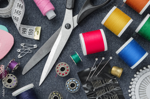 Sewing items: tailoring scissors, measuring tape, thimble, spools of thread, including pins, needles and sewing accessories on sewing cloth. View from above.