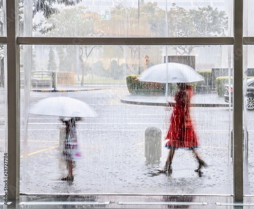 A tropical monsoon rain in Singapore viewed through a window streaked with water, as a woman and young girl walk outside carrying matching white umbrellas, their movement blurred.