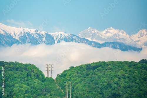Dense green forest in the valley and on the slopes of the mountain. Power lines in the forest. Snow capped mountains visible on the horizon